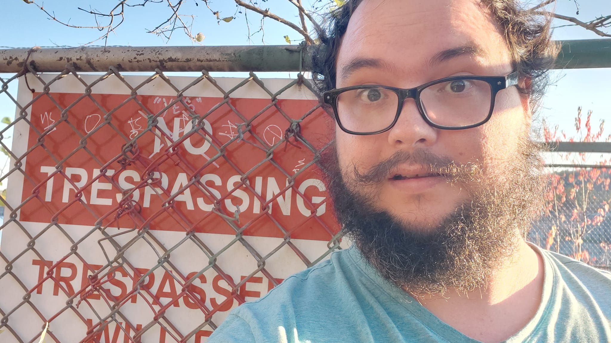 A man with short hair and bushy facial hair looks alarmed in front of a chain link fence and a sign that reads "No Trespassing, Trespassers will be Prosecuted"